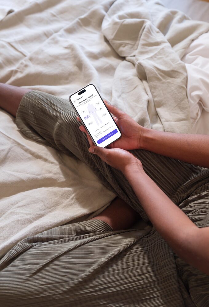 A woman sitting on a bed with the app’s injection map open on the phone in her hand, ready to log the dose