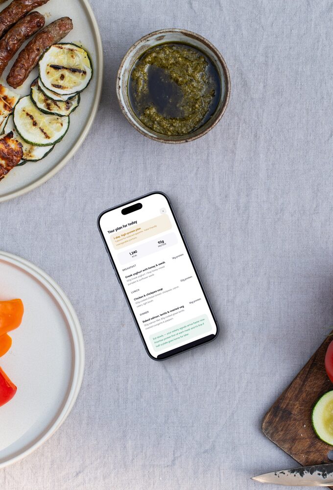 A table laid with grilled vegetables, halloumi and a bowl of dressing, the app’s day plan open on a phone beside them