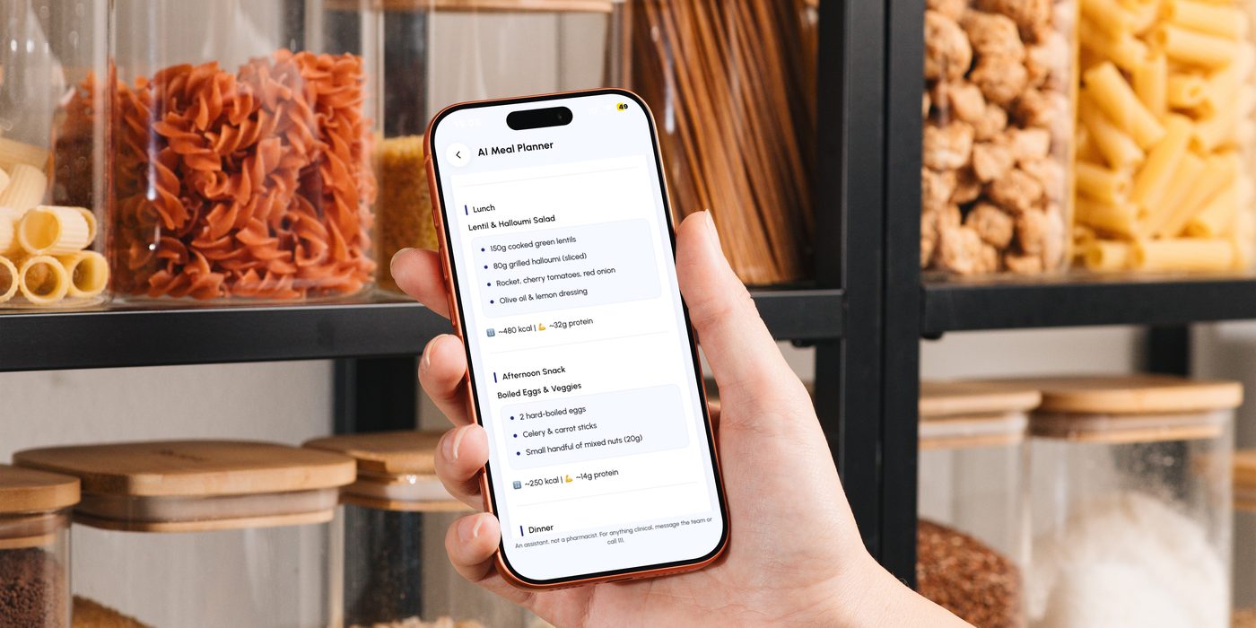 A phone held up in a kitchen with the app’s meal planner open, shelves of jars behind it